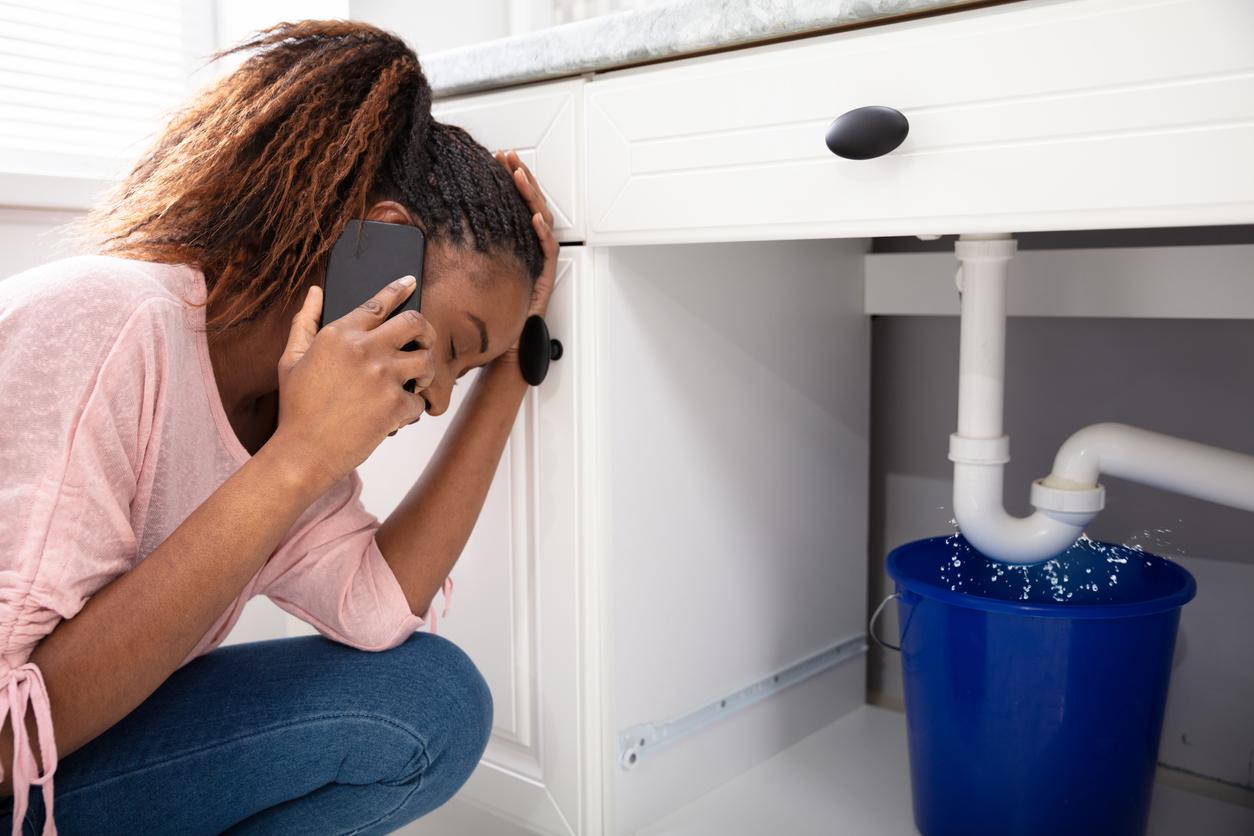 Young woman calling a plumber while crouching in front of a leaking sink pipe.